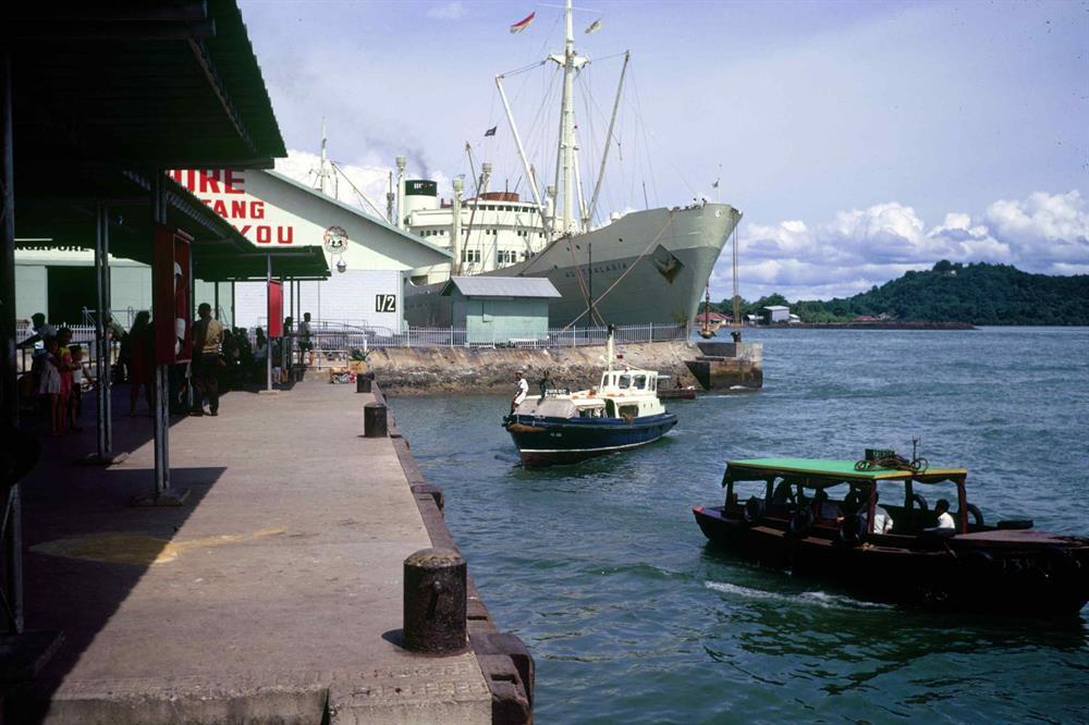 Large ship docked at a busy pier with people, smaller boats in the water, and "SINGAPORE THANK YOU" on a building.