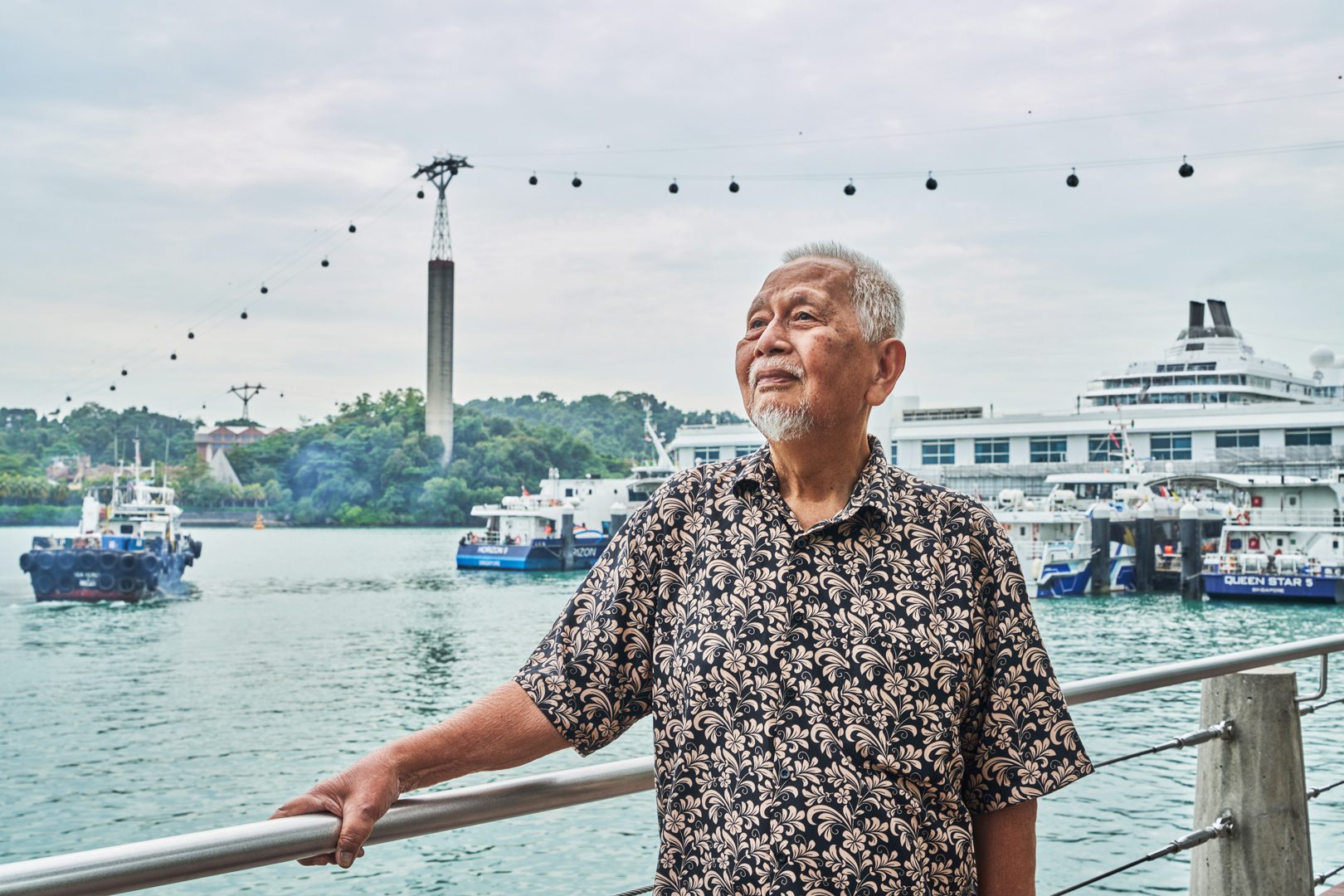 Elderly man by a waterfront with boats and a cable car system in the background.