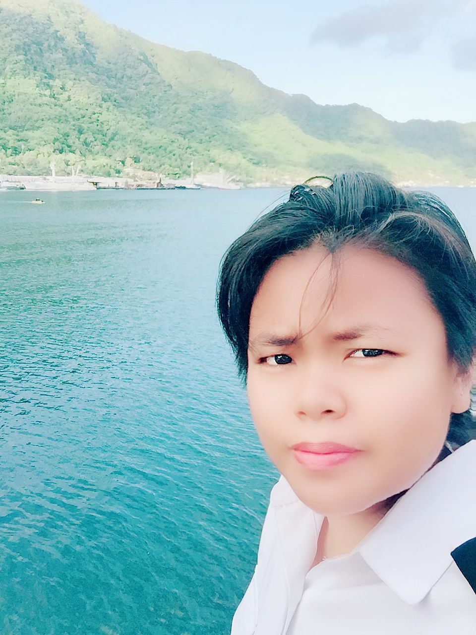 Young person in white shirt by ocean with green mountains.
