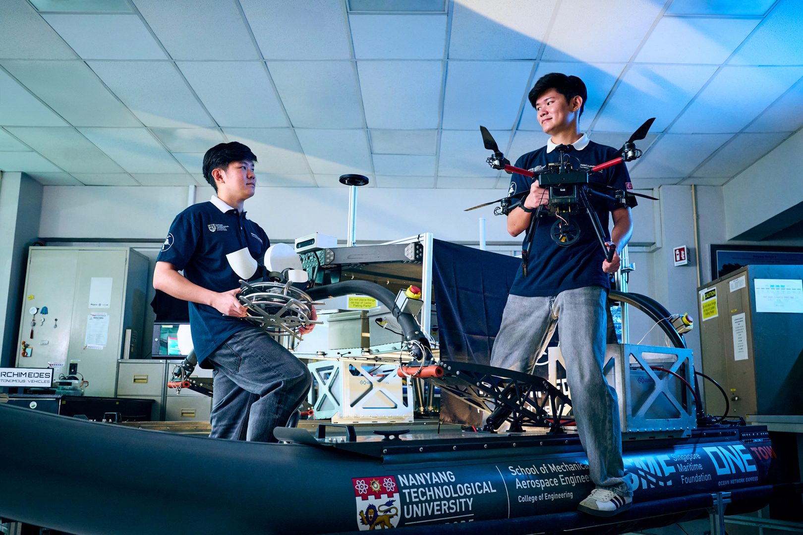 Two NTU engineering students in a lab, one holding a drone, near a maritime prototype.