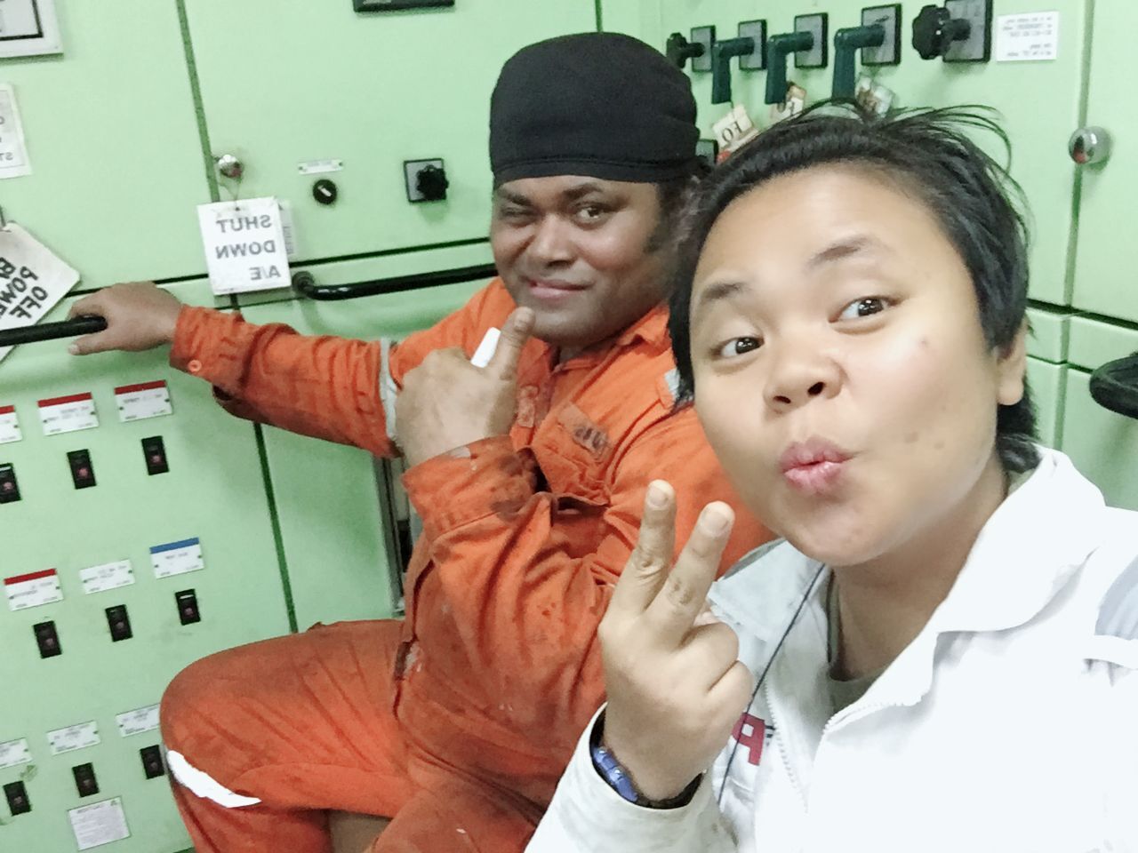 Two crew members pose in a control room, one giving a thumbs up, the other a peace sign.